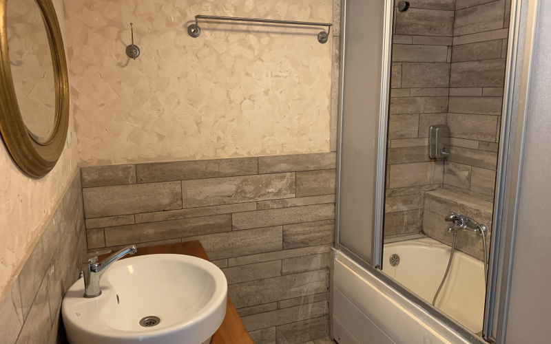 Banyo: küvet ve duş camı, ahşap görünümlü karolar, beyaz lavabo ve yuvarlak altın çerçeveli ayna.