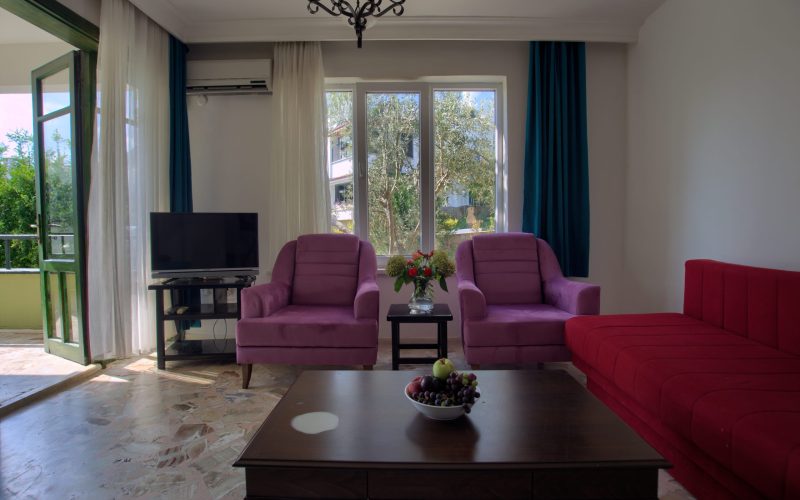 Salonun pencere tarafı: iki mor koltuk, kırmızı köşe kanepe, ortada ahşap sehpa ve meyve tabağı; camlı kapı balkona açılıyor, duvarda klima.