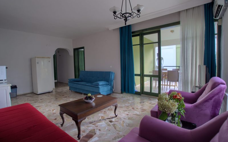 Geniş salon: mavi çekyat, kırmızı kanepe ve mor koltuklar; sürgülü camlı kapıdan üstü kapalı balkondaki yemek masası görünüyor, solda buzdolabı.