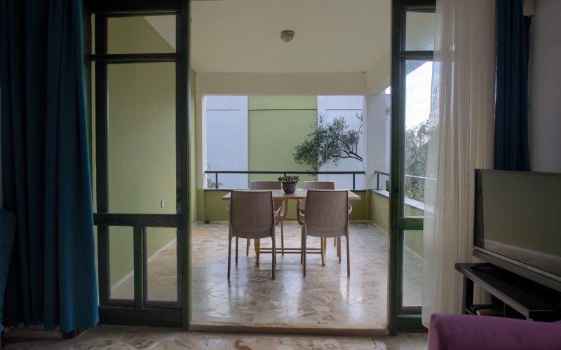 Salondan balkona bakış: açık camlı kapının ardında yeşil duvarlı üstü kapalı balkon, dört sandalyeli yemek masası ve zeytin dalları.