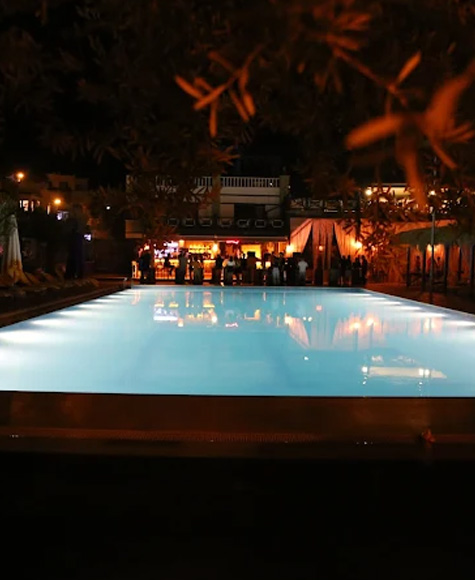 Gece havuz başı: aydınlatılmış havuzun ardında kalabalık bir bar, sıcak ışıklar ve ağaçların silueti.