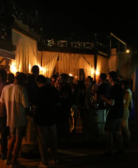 Gece açık havada parti: bar önünde ayakta sohbet eden kalabalık, arkada beyaz perdeler ve sıcak sarı aydınlatma.