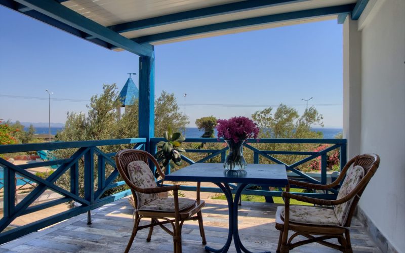Üstü kapalı balkon: mavi ahşap korkuluk, mavi masa ve iki hasır koltuk, masada vazoda mor çiçekler; arkada zeytin ağaçları ve deniz.